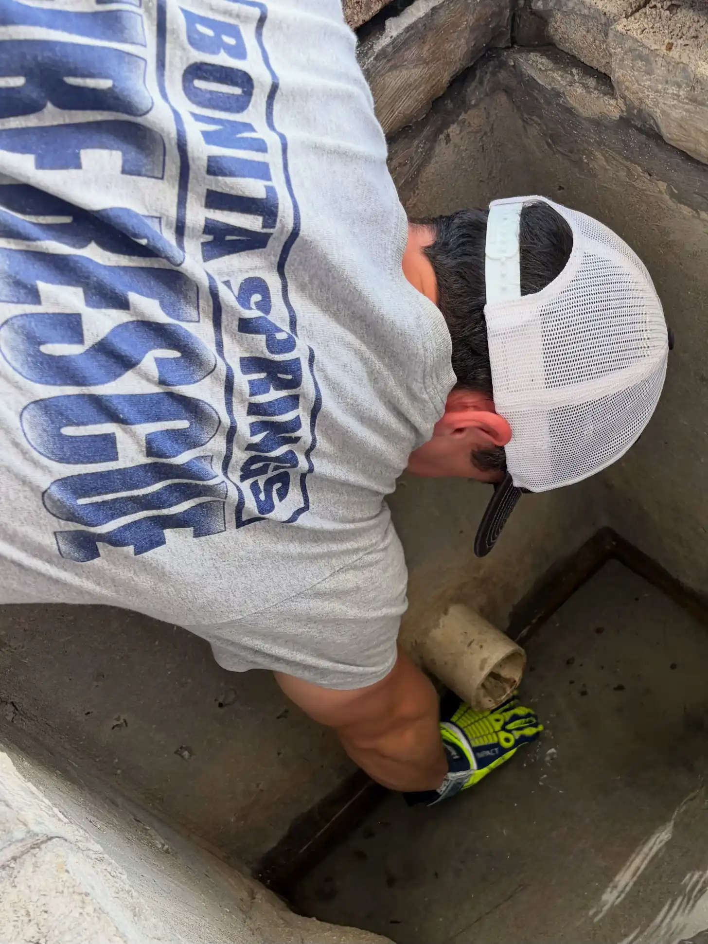 A person wearing a white cap and blue and white graphic t-shirt stands in a concrete drainage channel, wearing neon yellow and black sneakers.