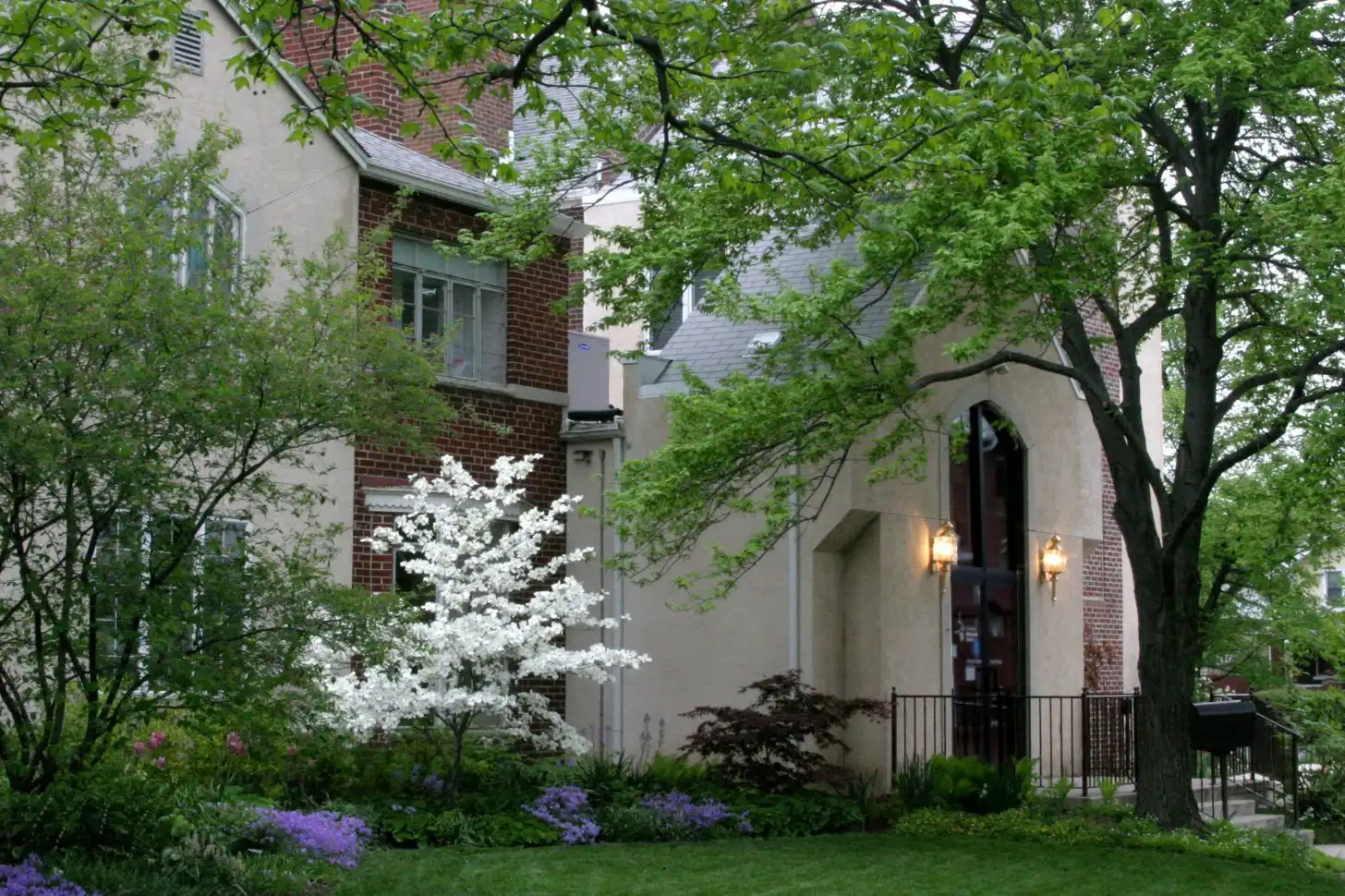 Brick and stone residential building with white flowering shrub and purple flowers in front garden, surrounded by leafy trees.