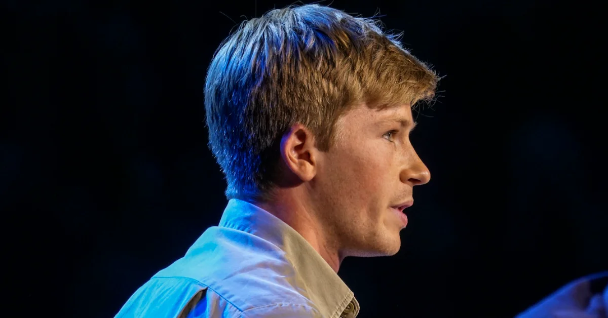 Man in profile wearing a light blue shirt against a dark background, illuminated by blue stage lighting.