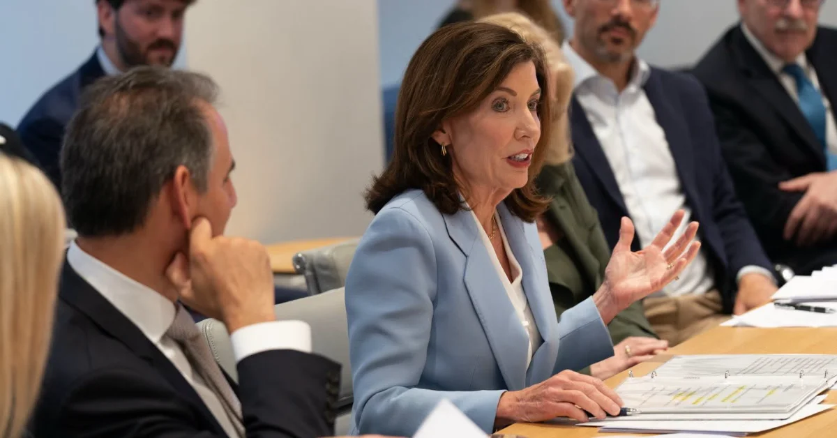 Woman in light blue blazer gestures while speaking at a table during a meeting with multiple people seated and standing around her. Documents are visible on the table in front of her.
