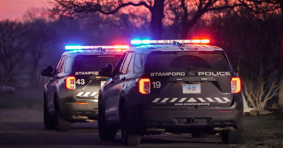 Two Stamford Police vehicles with emergency lights activated parked on a road at dusk, with bare trees visible in the background.