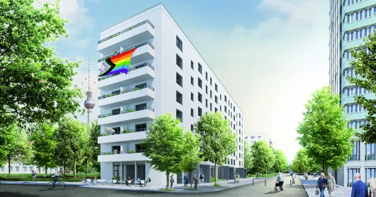 White modern apartment building with a rainbow flag on its facade, situated among green trees and other residential buildings in an urban street scene.