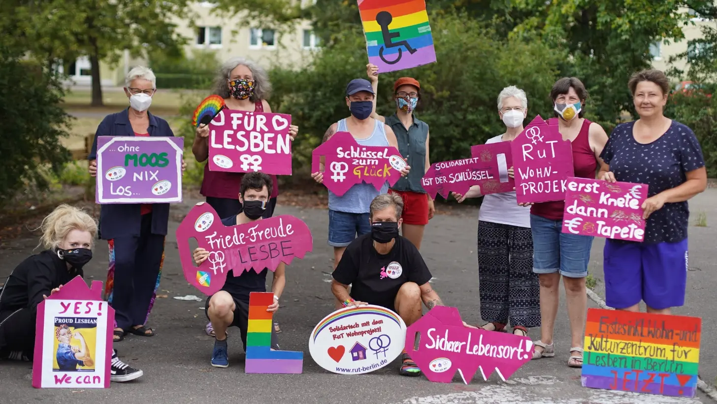 A group of people holding rainbow-colored signs with German text stand together on a street. Signs read "FÜR LESBEN" (For Lesbians), "Friede-Freude LESBE," "Schlüssel zum Glück," and "Sicher Lebensort!" One person holds a rainbow disability pride flag.