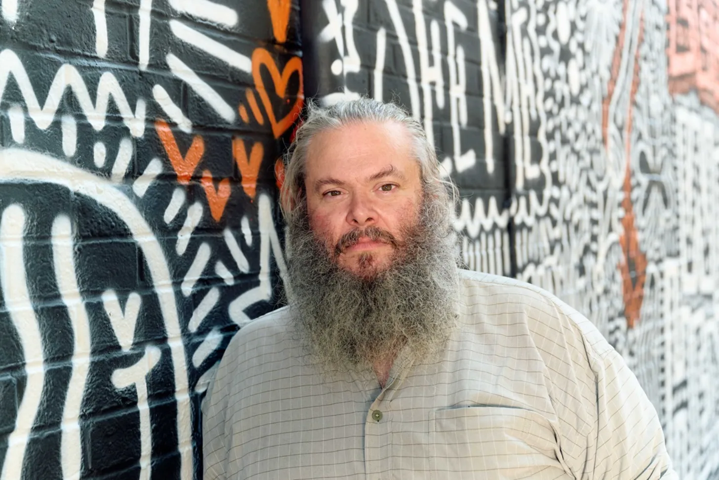 Man with long gray beard and gray hair wearing a tan button-up shirt stands in front of a brick wall with black and white graffiti and orange accents.