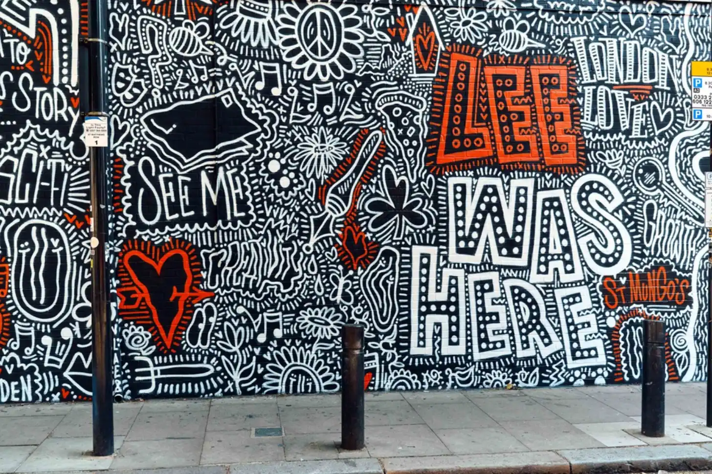 Street mural with black and white intricate patterns featuring the text "LIFE WAS HERE" in large white letters with dot patterns, "Lee was here," in red letters at top, and a red heart shape. Dark bollards in foreground on paved sidewalk.