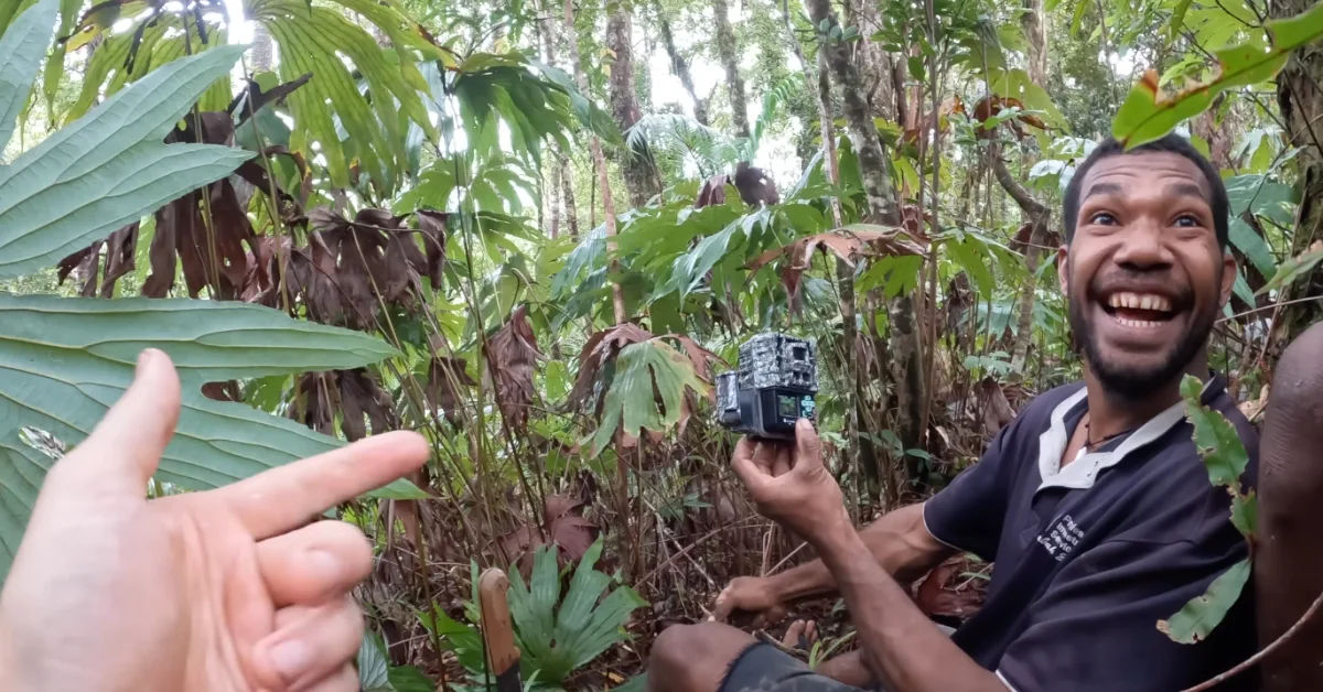 A man in a dark shirt holds a small electronic device and smiles at the camera in a dense tropical forest with green and brown vegetation surrounding him.