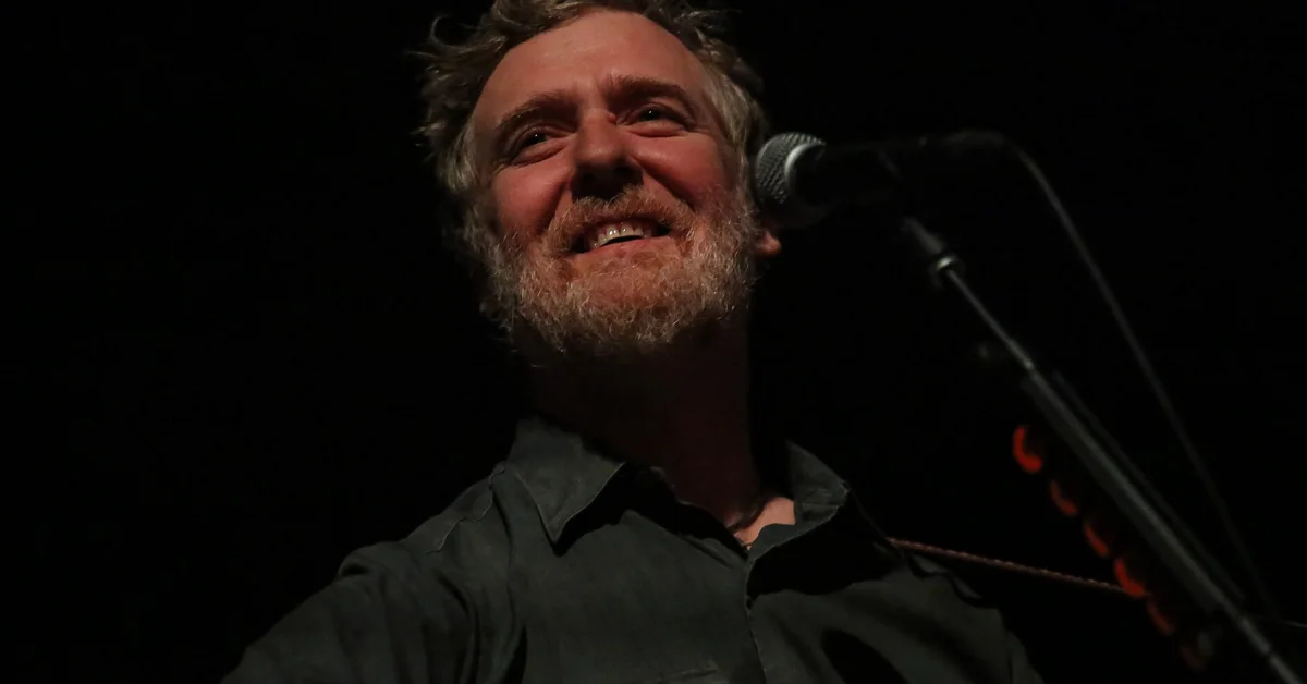Glen Hansard pictured: A man with a beard wearing a dark shirt speaks at a microphone against a black background.