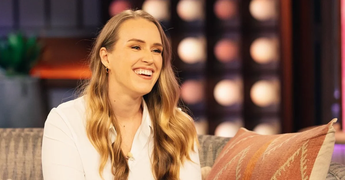Woman with long blonde hair wearing a white blouse, smiling while seated on a gray couch with an orange and cream striped pillow, against a blurred background with warm lighting.