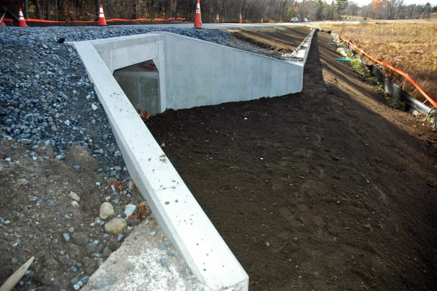Concrete culvert structure under construction with white walls and dark soil interior, surrounded by gravel and construction barriers with traffic cones.