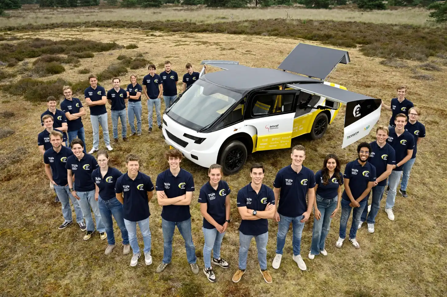 A white and yellow car with open doors and roof panels stands in a field surrounded by approximately 30 people wearing matching dark navy polo shirts with logos. The vehicle displays branding on its side.