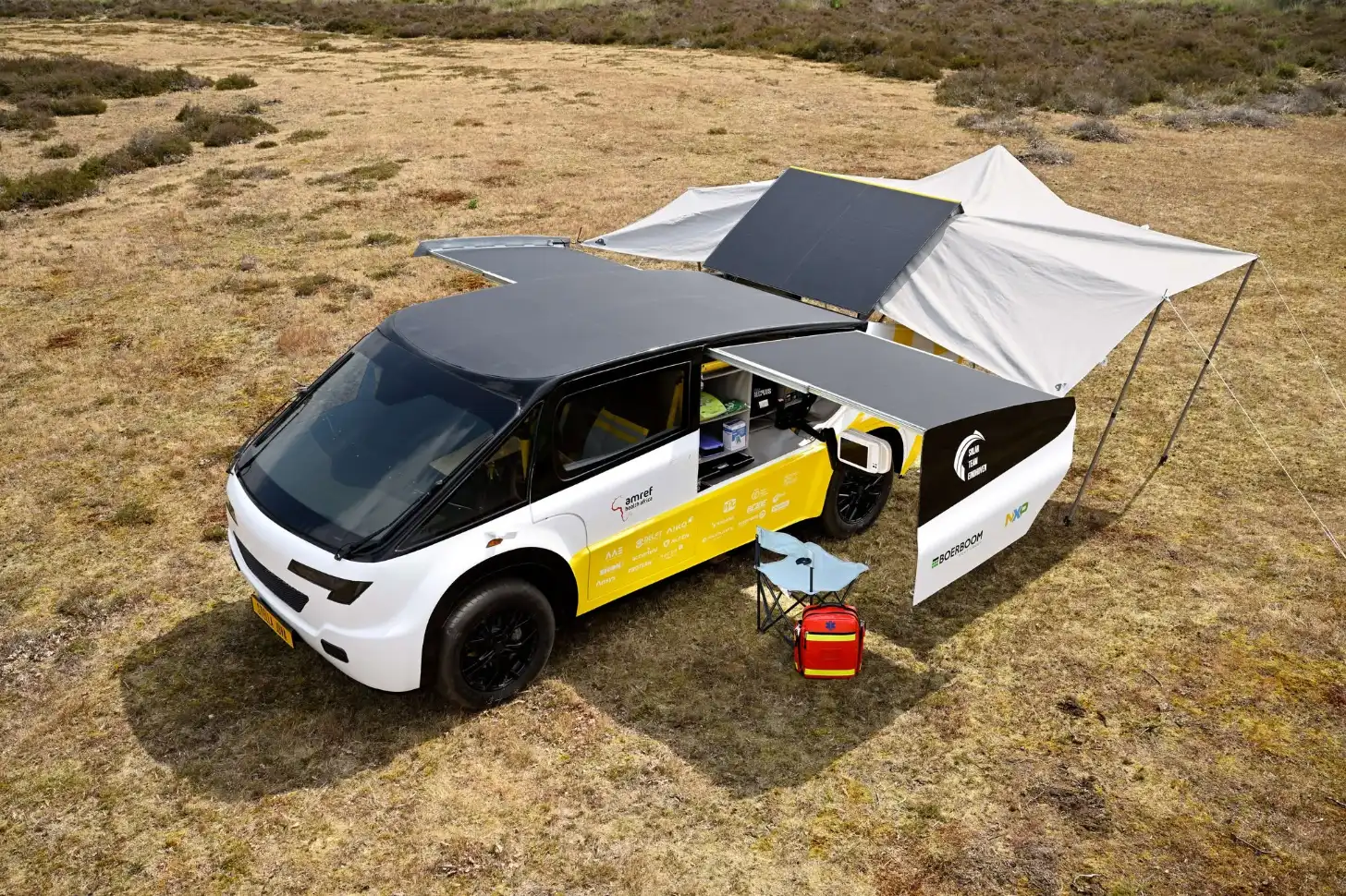 A white and yellow vehicle with an open door is parked in a desert landscape with multiple black and white sun shade panels extended overhead. A red container and camping chair sit nearby on the ground.