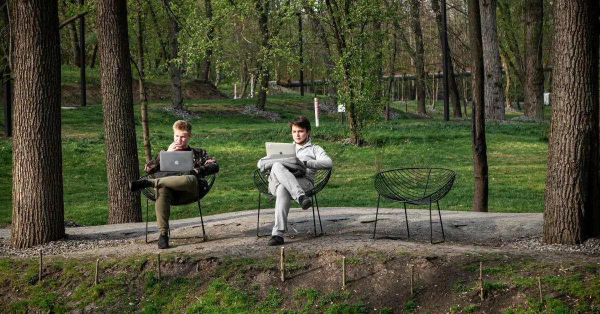 Two people sit in chairs in a wooded area, each working on a laptop computer.