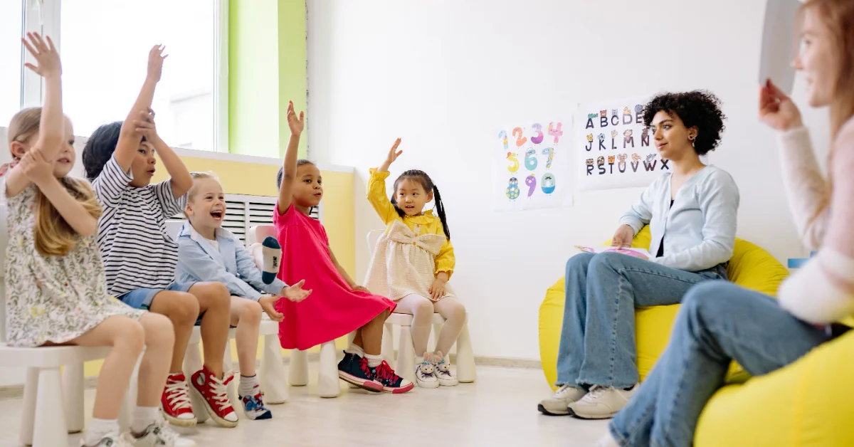 A teacher sitting on a yellow chair speaks to a group of young children seated in front of her in a classroom with alphabet and number posters on the wall. Several children have their hands raised.