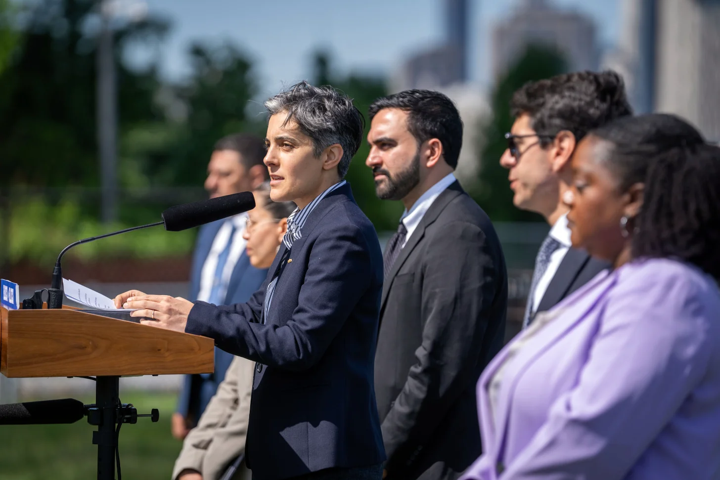 A man in a dark blazer speaks at a wooden podium with a microphone, holding papers. Several other people in business attire stand beside him outdoors, with buildings and greenery visible in the background.