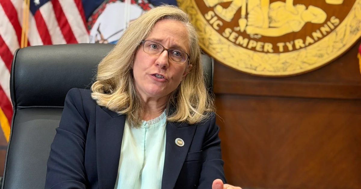 A woman, Virginia Governor Abigail Spanberger, wearing glasses and dark blazer seated at desk speaking, with American flag and gold seal visible in background.