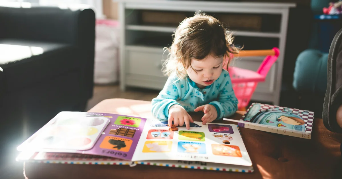 A toddler in a teal patterned shirt looks down at an open picture book with colorful illustrated pages on a table in a kitchen.