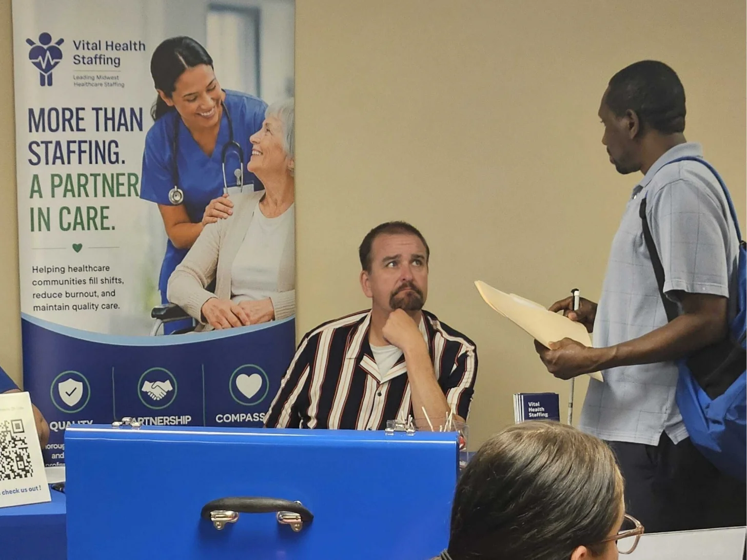A man in a striped shirt sits at a blue table with a Vital Health Staffing banner behind him, speaking with a man in a light blue shirt who holds papers. A woman with gray hair sits in the foreground. The banner reads "MORE THAN STAFFING. A PARTNER IN CARE."
