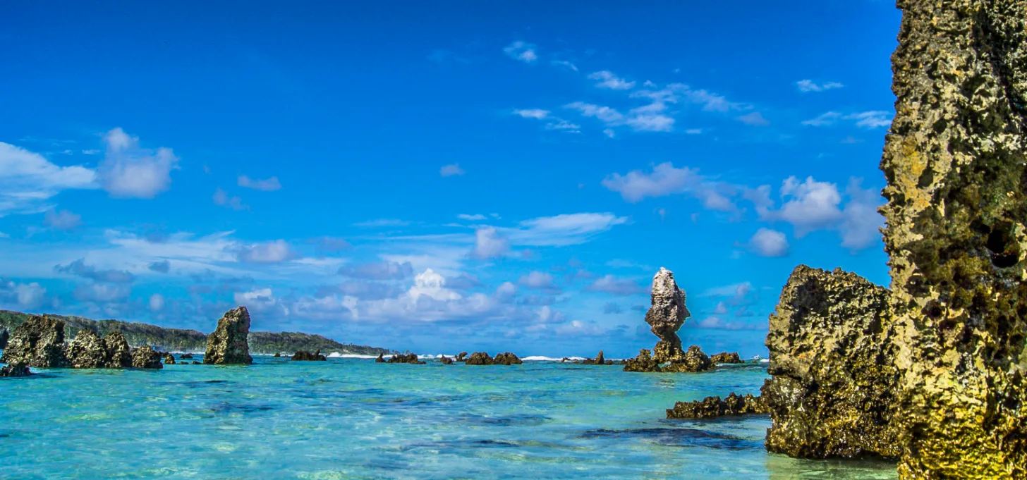 Turquoise water with lichen-covered rock formations in the foreground and distant rocky outcrops under a blue sky with white clouds.