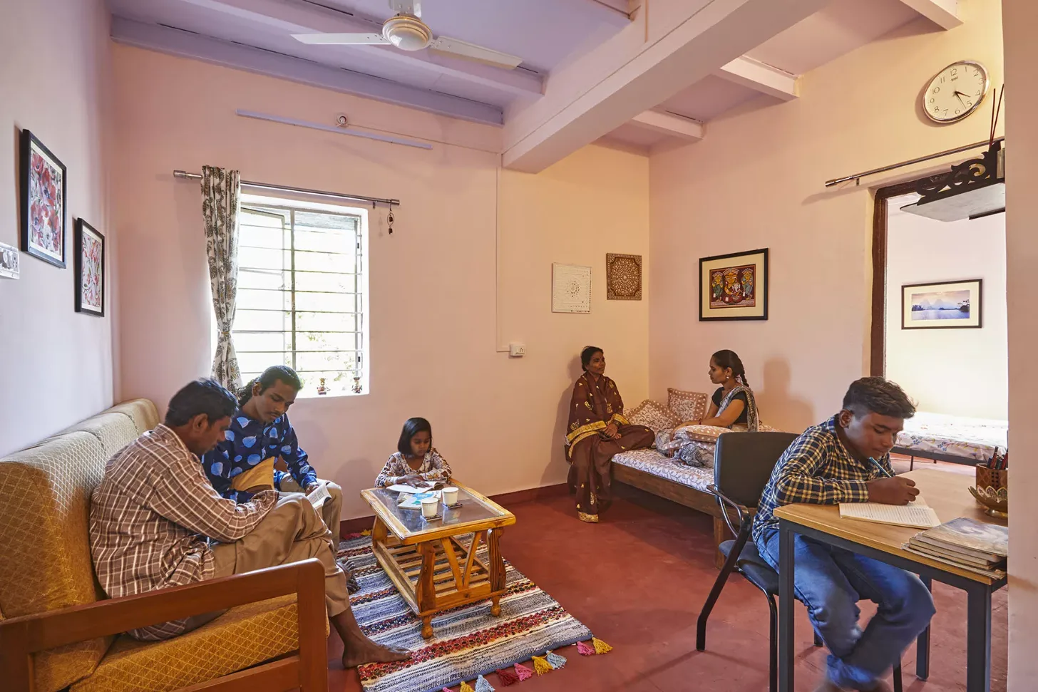 Several people sit in a peach-colored room with red tile flooring. Two men on the left read from papers while seated on a wooden bench; a girl sits at a small wooden table in the center; two women in traditional dress sit on a bed in the background; a man at a desk on the right writes in a notebook. A wall clock, framed artwork, and a window with patterned curtains are visible.