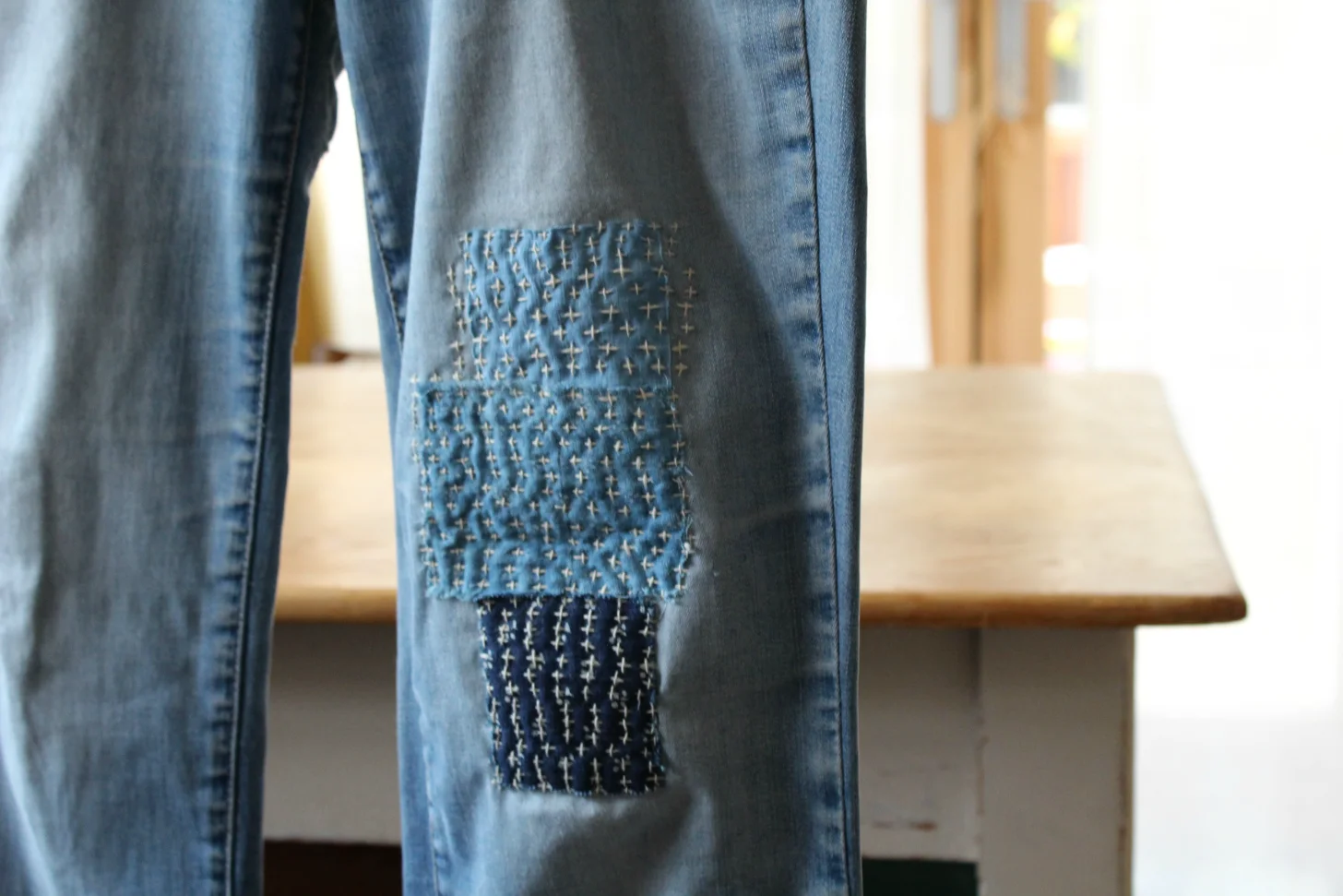 Jeans with decorative cross-stitch patches on the thigh, displayed on a wooden bench indoors.