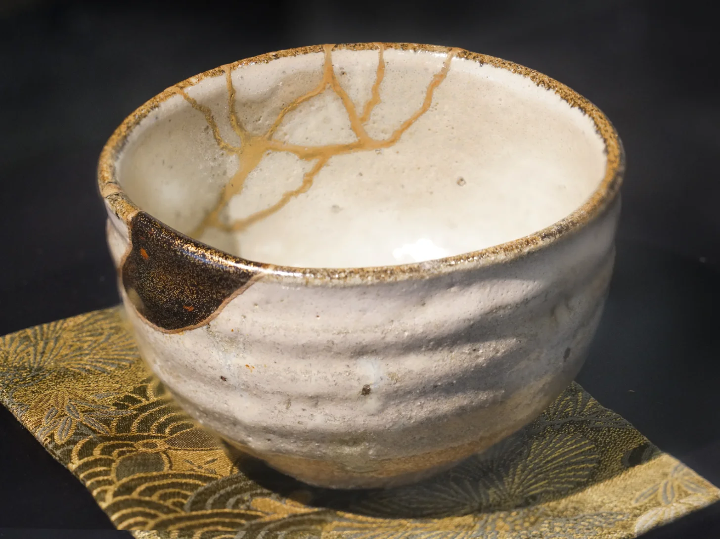 A ceramic bowl with a cream-colored glaze sits on a decorative gold and dark patterned cloth. The bowl features brown brushstroke designs on its interior rim and exterior surface, with a rough, textured finish around its edges.