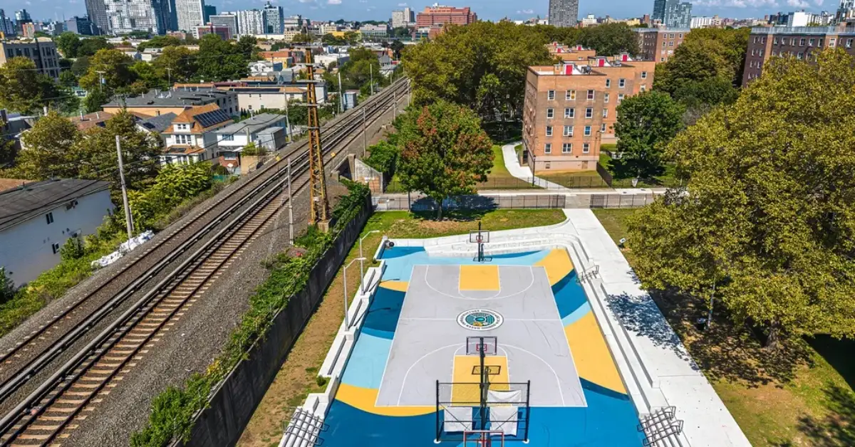Aerial view of a blue and yellow basketball court with multiple hoops surrounded by brick apartment buildings, railroad tracks, and trees in an urban neighborhood with a city skyline in the distance.