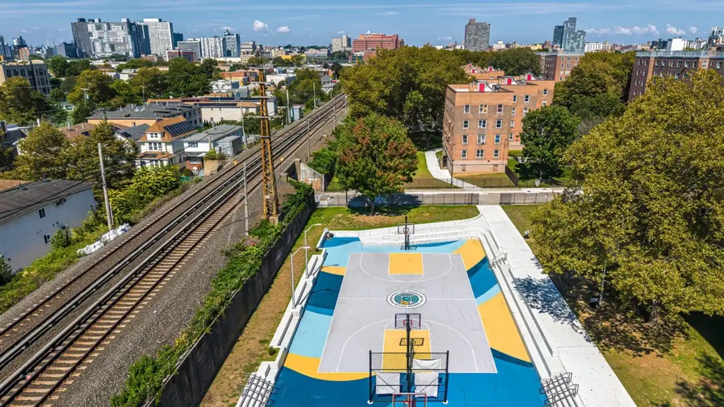 Aerial view of a blue and yellow basketball court surrounded by brick apartment buildings, railroad tracks, and green trees in an urban neighborhood with a city skyline visible in the distance.