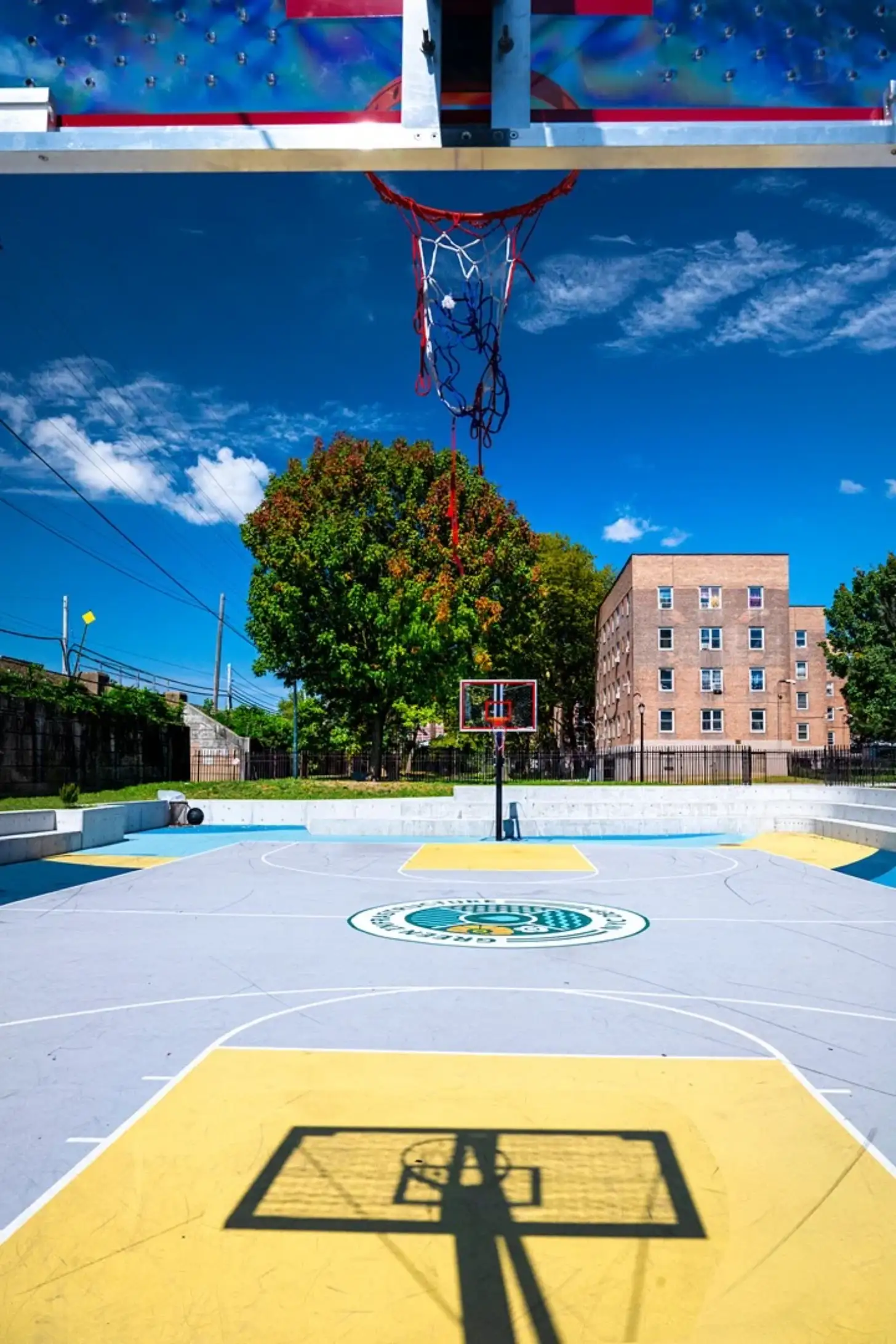 An outdoor basketball court with yellow and blue sections, a center court logo, and a basketball hoop with red net. A large tree, brick apartment building, and clear blue sky are visible in the background.