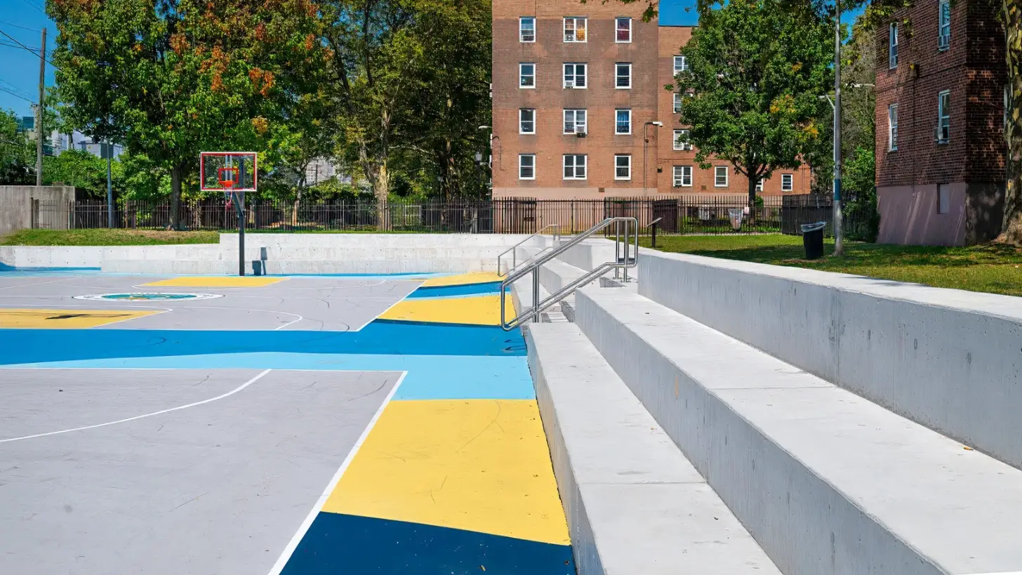 A colorful outdoor sports court with blue and yellow painted sections sits below a concrete bleacher structure. Brick residential buildings frame the tree-lined urban playground in the background.