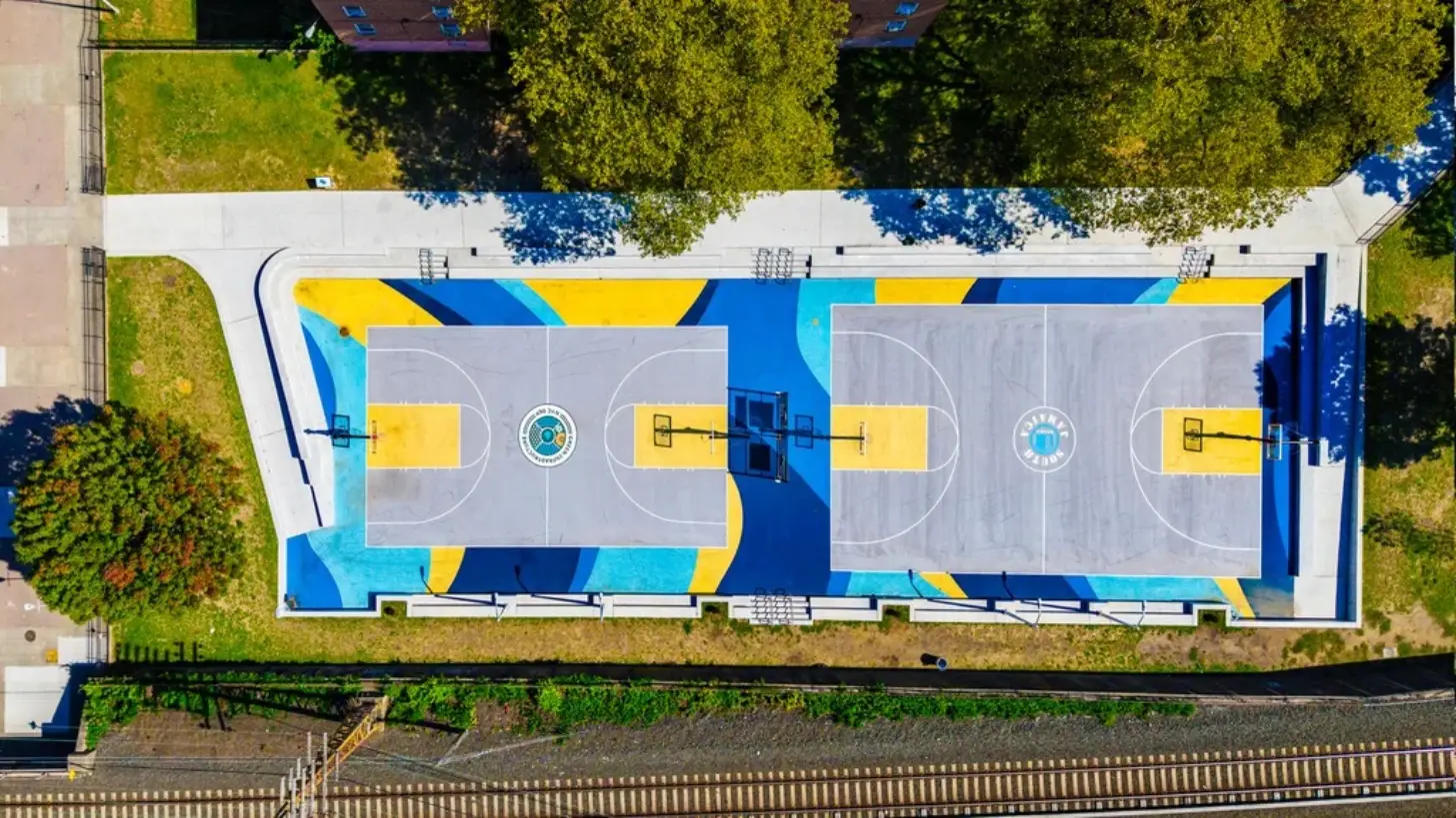 Aerial view of two indoor basketball courts with gray playing surfaces, yellow end zones, and blue and yellow border designs. Blue basketball hoops are mounted above each court. Surrounding areas show manicured grass and trees.