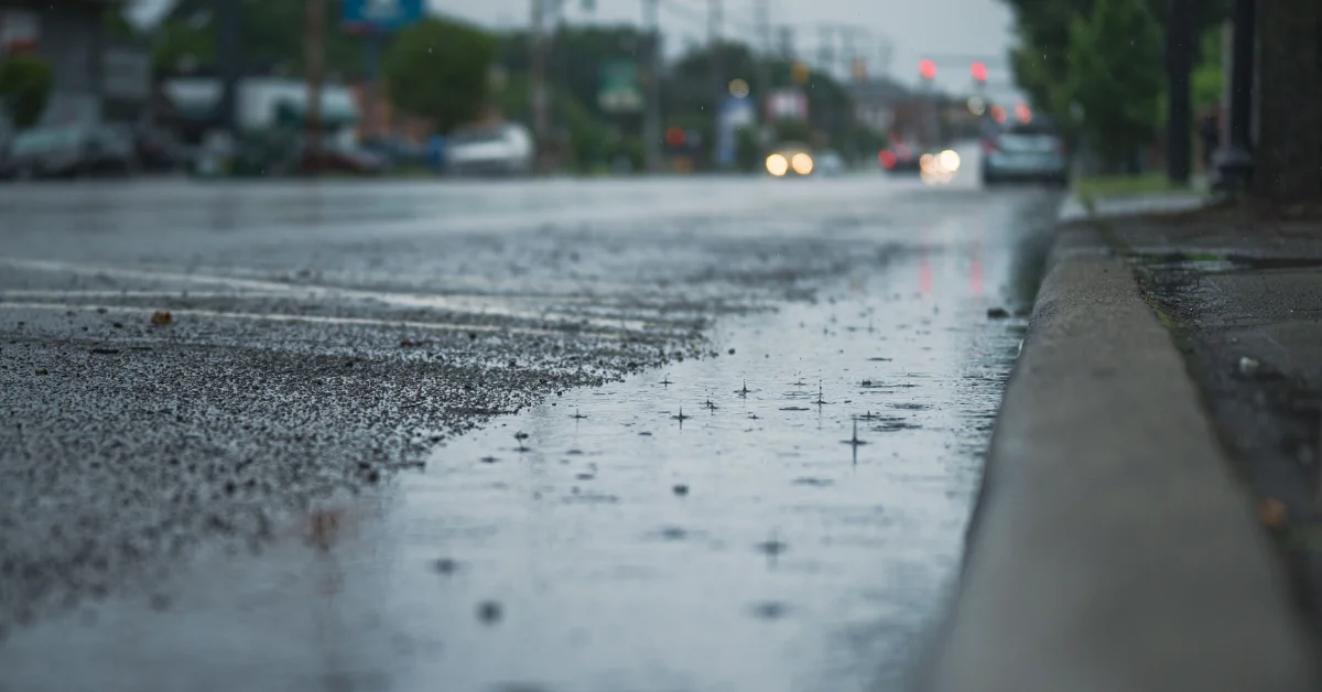 Close-up of a wet sidewalk with raindrops visible on its surface, blurred vehicles and street lights in the background.