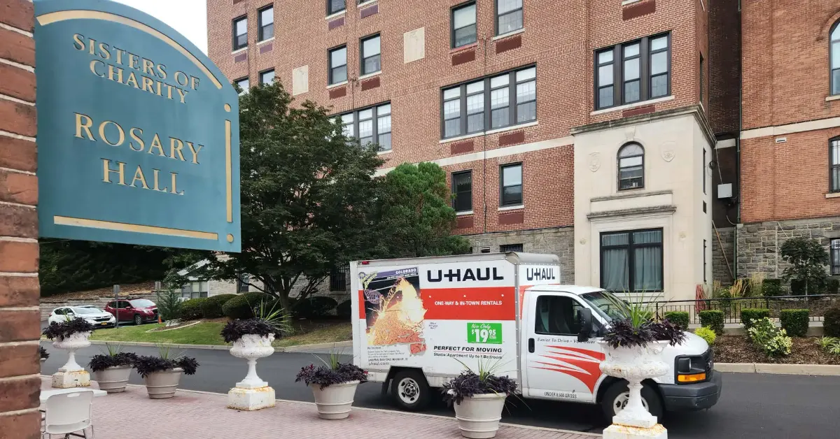 U-Haul rental trucks parked in front of Rosary Hall, a brick building operated by Sisters of Charity, with landscaped planters in the foreground.