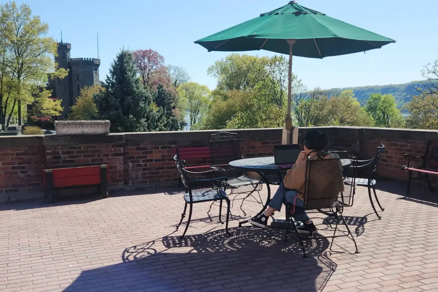 Person sitting at a table on a brick terrace with green umbrella, castle tower visible in background, river and forested hills beyond.