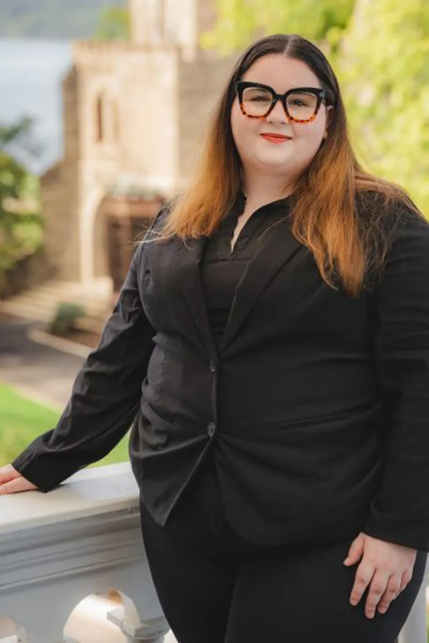 Woman wearing black blazer and tortoiseshell glasses stands outdoors against a blurred stone building and green foliage background.