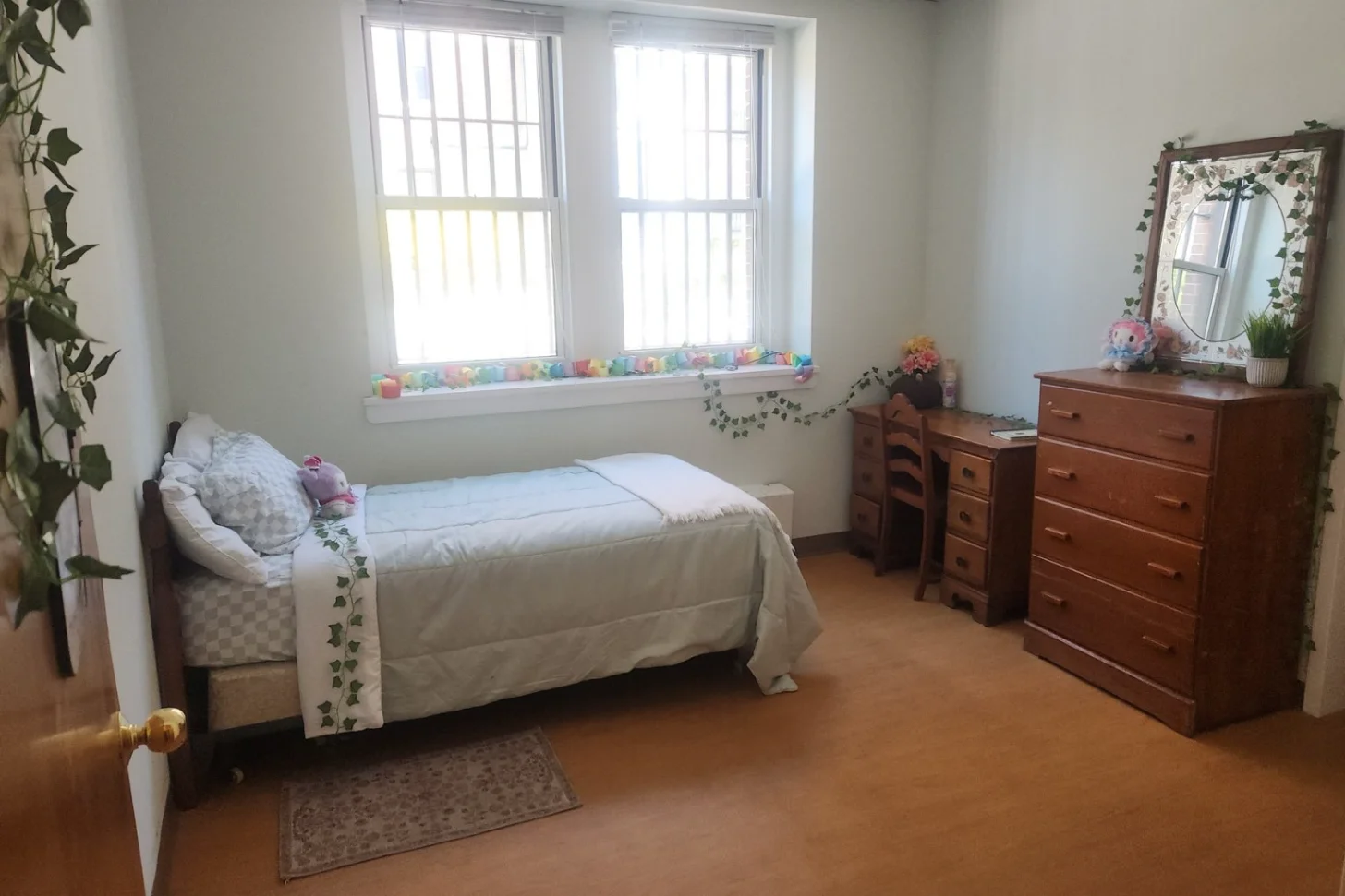 A bedroom with a single bed, wooden dresser, desk, and mirror. Two windows with colorful objects on the windowsill overlook the room. Plants decorate the space, and hardwood flooring covers the floor.
