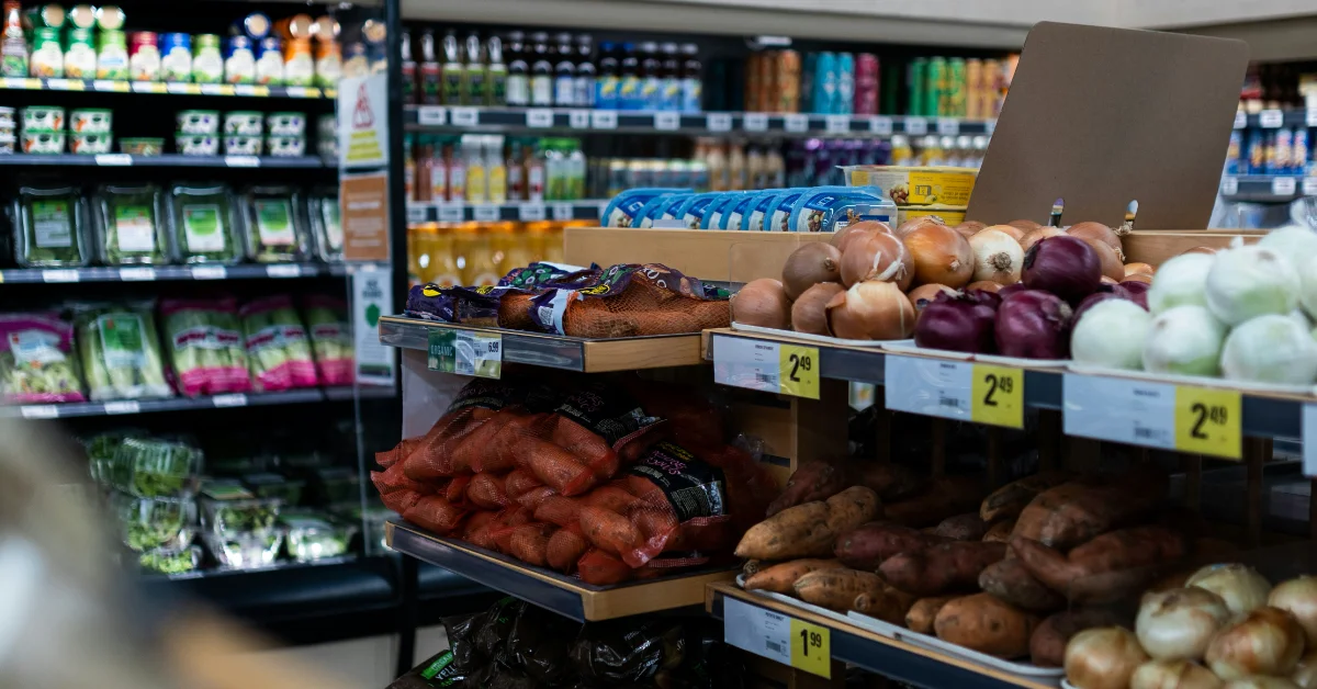 Grocery store produce section with display bins containing potatoes, yams, and yellow, white, and purple onions, priced at $1.99 and $2.99. Refrigerated cases and shelves stocked with beverages visible in the blurred background.
