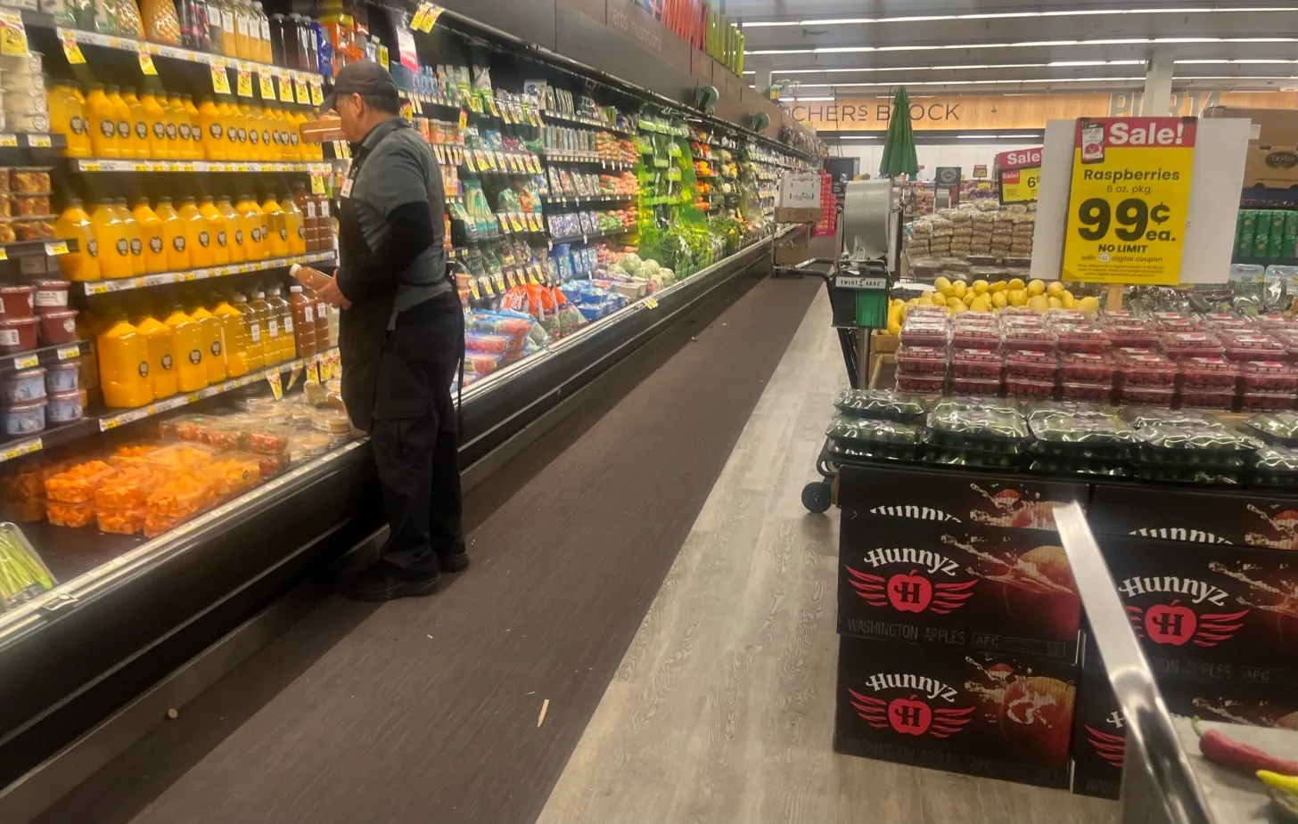 A person in dark clothing stands at a refrigerated produce section in a grocery store, with yellow juice bottles on the left shelf and fresh vegetables visible behind glass. On the right, yellow sale signs advertise raspberries for 99 cents, with packaged produce and boxes of apples displayed below.