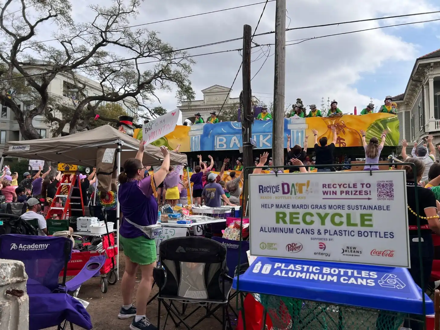 A recycling collection booth at a Mardi Gras parade displays a sign reading "Recycle Dat! Recycle to Win Prizes! Help Make Mardi Gras More Sustainable. Recycle Aluminum Cans & Plastic Bottles." Parade-goers in purple and gold clothing walk past floats and tents along a tree-lined street.