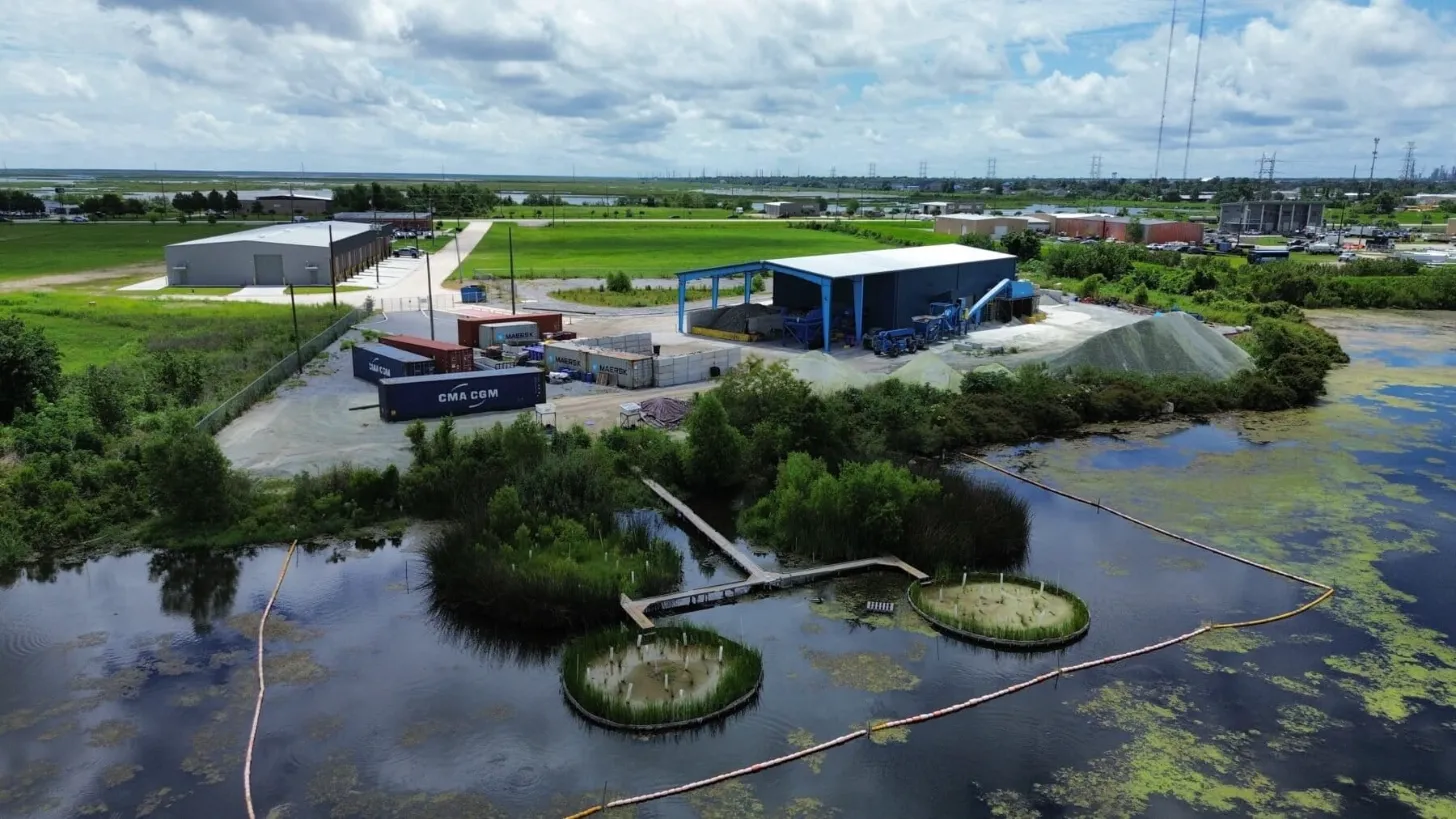 Aerial view of an industrial waterfront facility with shipping containers, blue-roofed warehouses, gray storage structures, and containment booms surrounding wetland areas with vegetation islands in the foreground.