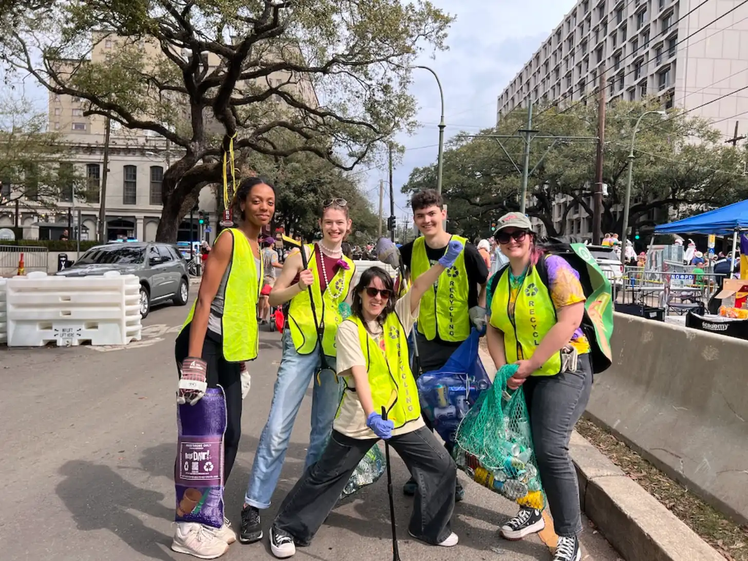 Five people wearing bright yellow safety vests stand together on a city street, smiling at the camera. One person holds a purple recycling bin and another holds a green mesh bag filled with items.