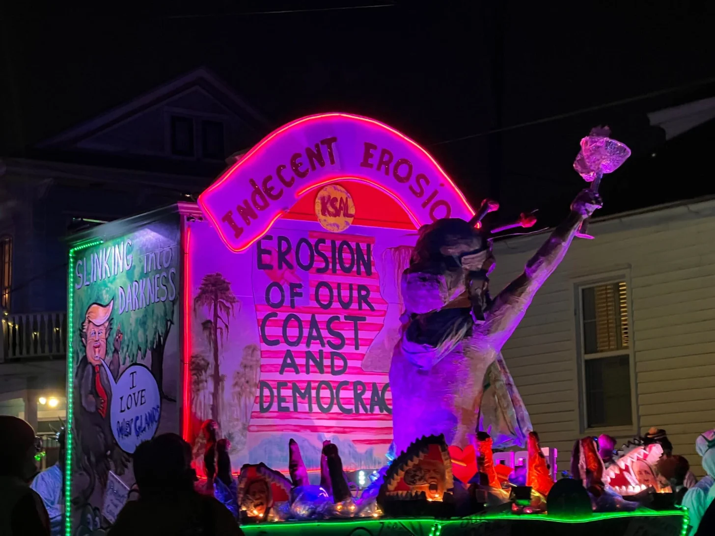 A neon sign shaped like a coastal erosion awareness monument displays text reading "Indecent Erosion of Our Coast and Democracy" in pink and red lighting, with a large sculptural figure raising its arm. A separate green neon sign reading "Sinking into Darkness" with a portrait is visible to the left.