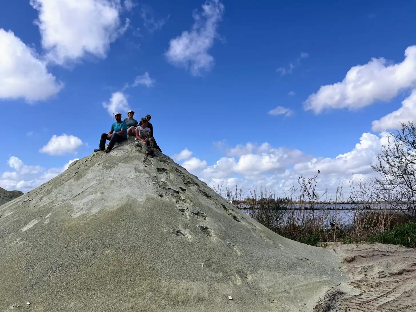 Three people sit atop a large conical mound of light gray sand against a blue sky with white clouds. A body of water with bare trees lines the background to the right.