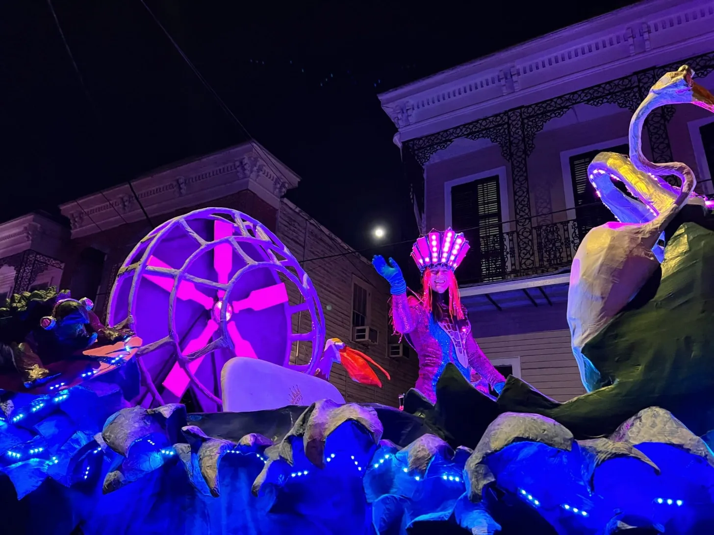 Nighttime outdoor display with neon-lit sculptural figures and structures in magenta, blue, and yellow colors in front of historic buildings with ornate architecture.