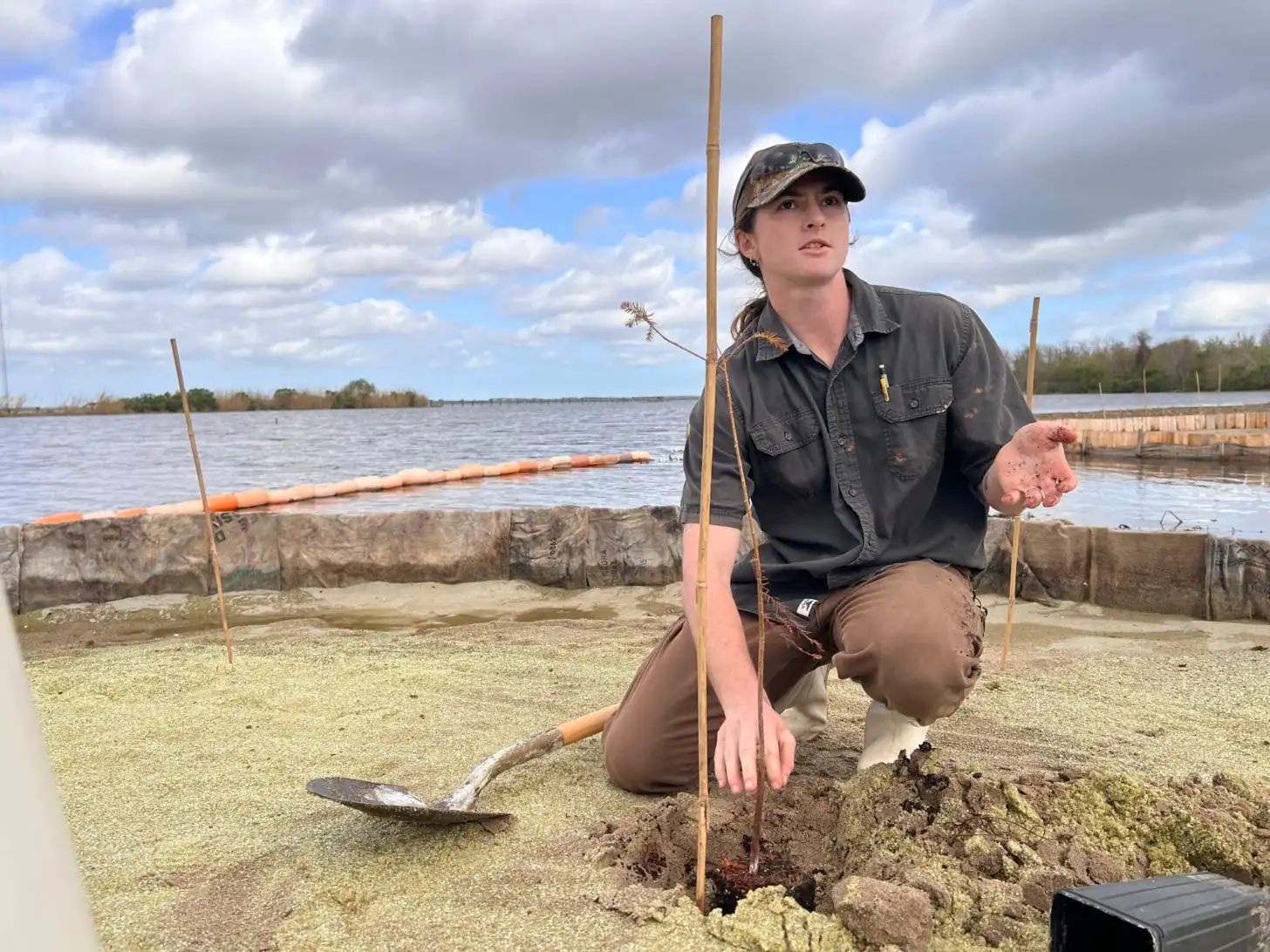 A person in a camouflage cap and dark shirt crouches beside a hole in sandy ground near a waterfront, holding soil in their hand with wooden poles and measuring tools visible nearby.