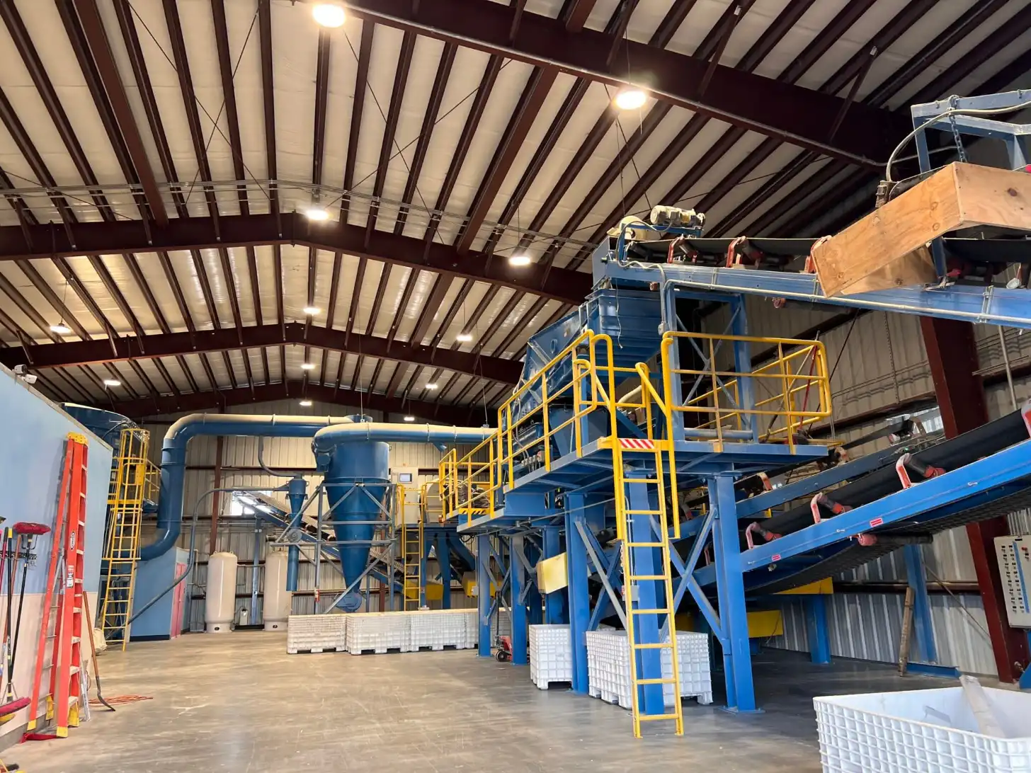 Industrial processing facility with blue steel structure, yellow safety railings and ladders, and large cylindrical equipment inside a metal warehouse with exposed beam ceiling and LED lighting.