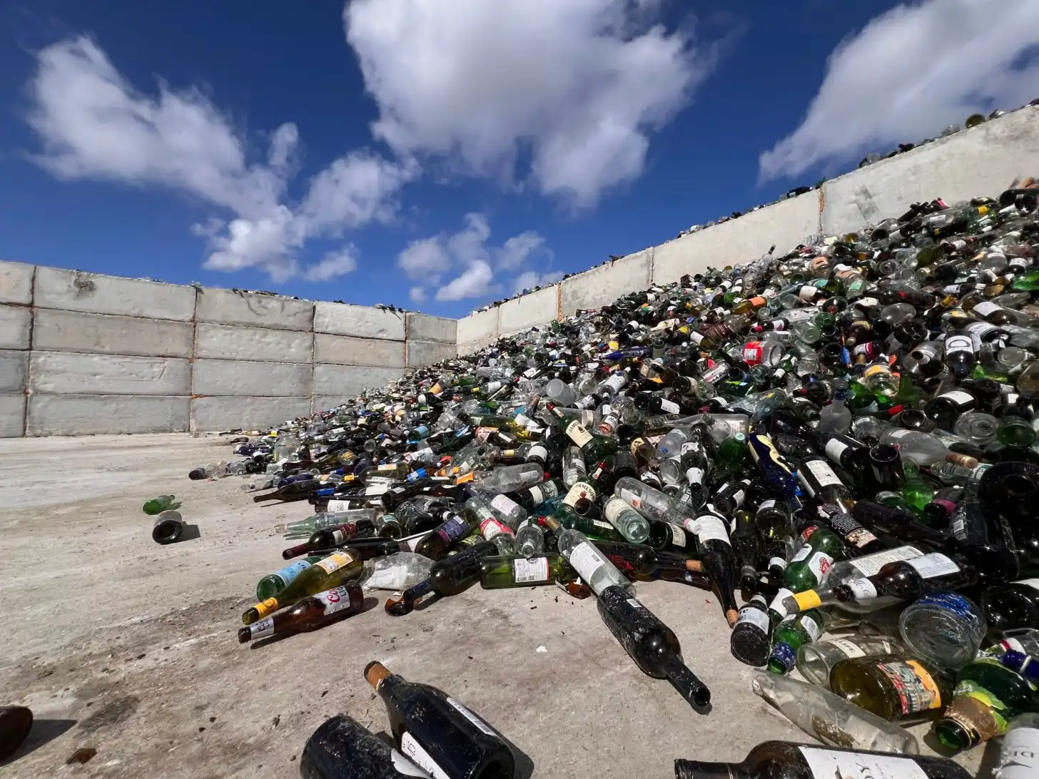 Thousands of glass bottles piled against concrete walls in an open-air facility under blue sky with white clouds.