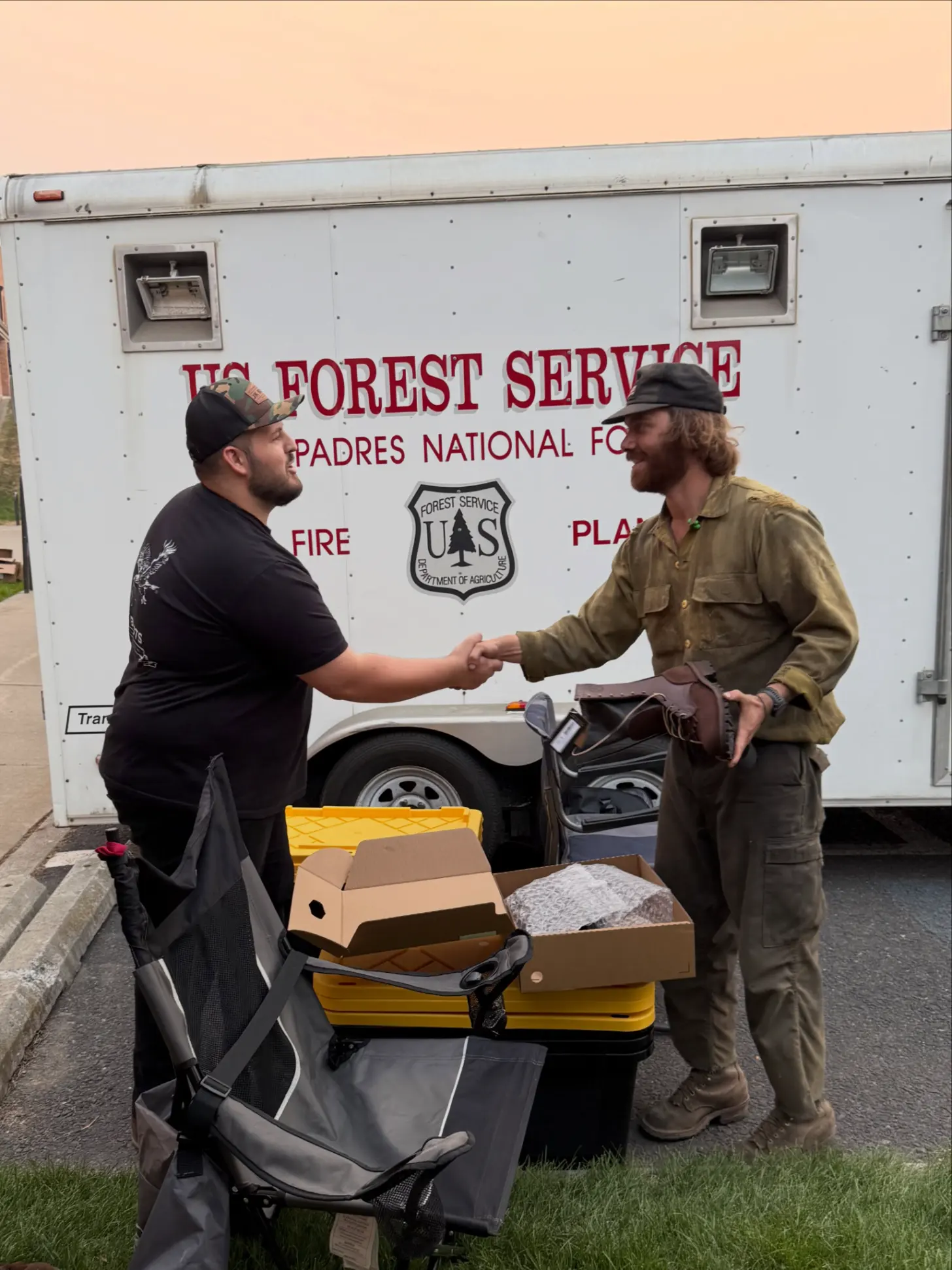 Two men shake hands in front of a U.S. Forest Service vehicle. Boxes and equipment are visible on the ground between them.