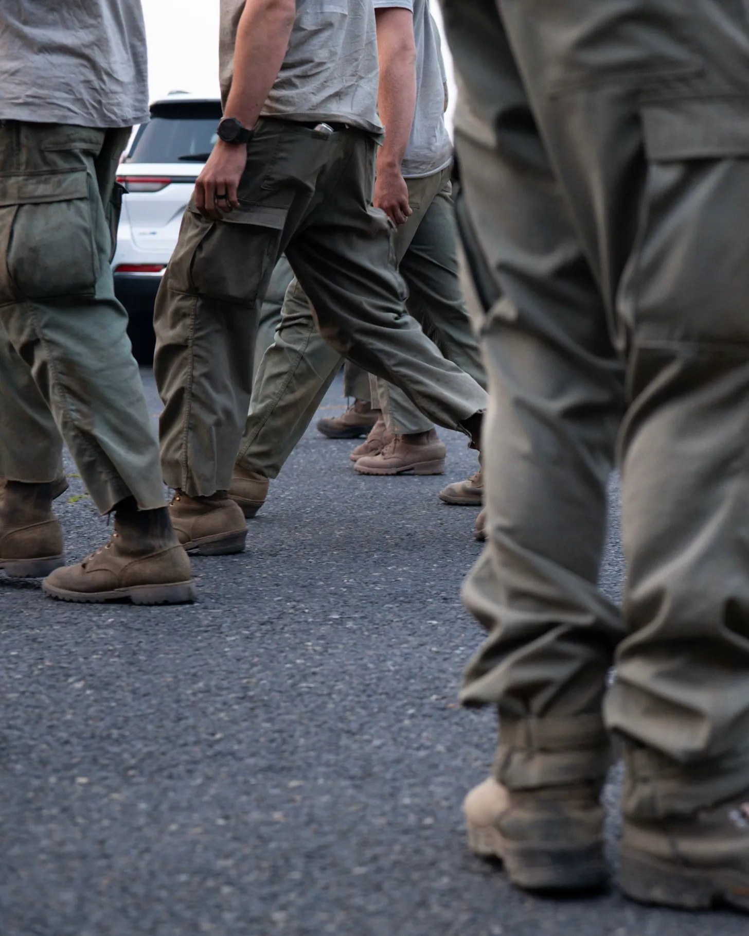 Multiple people wearing cargo pants and boots stand on pavement near parked vehicles.
