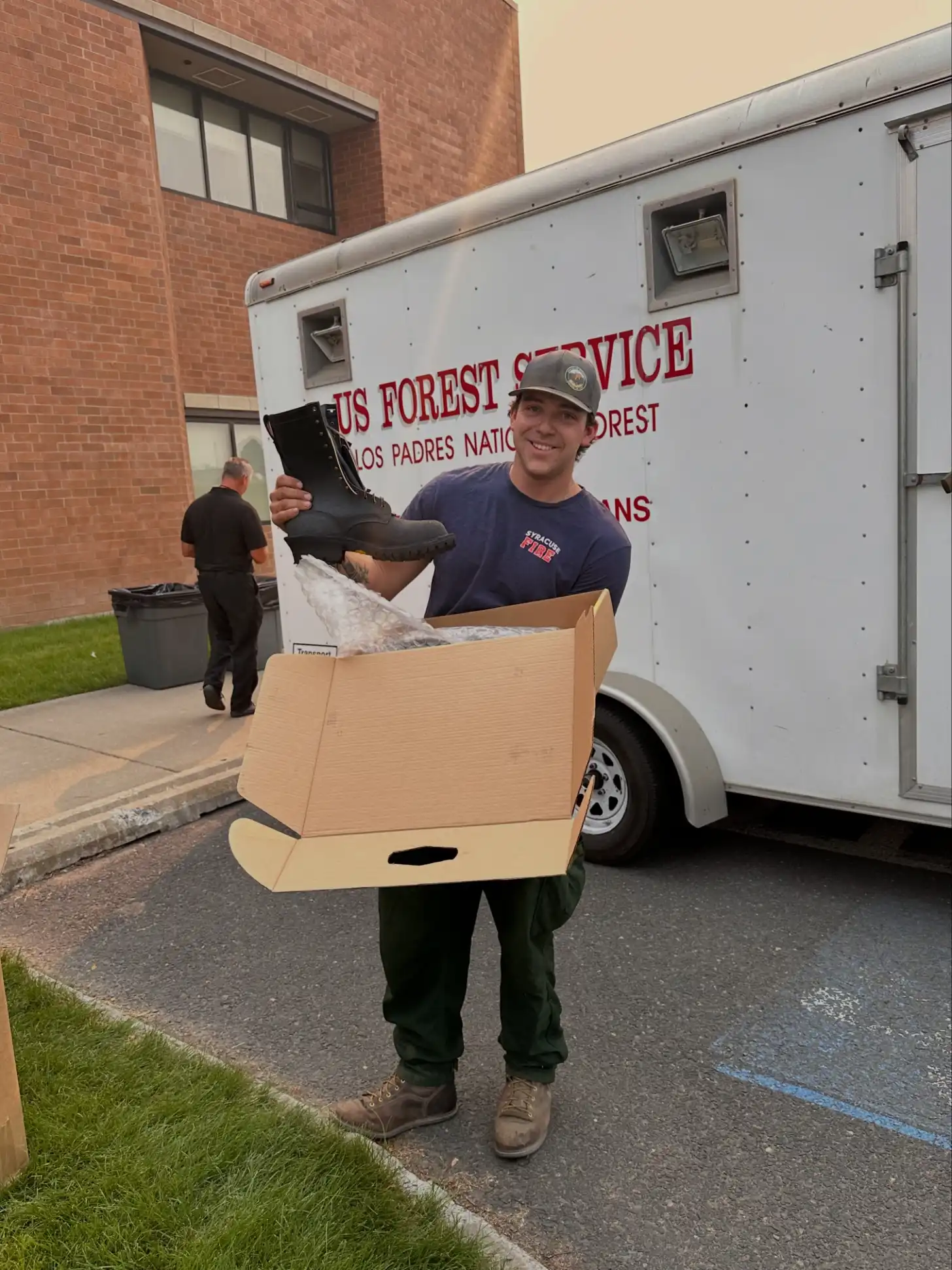 A U.S. Forest Service employee holds an open cardboard box containing a black hiking boot and other items in front of a Los Padres National Forest service trailer.