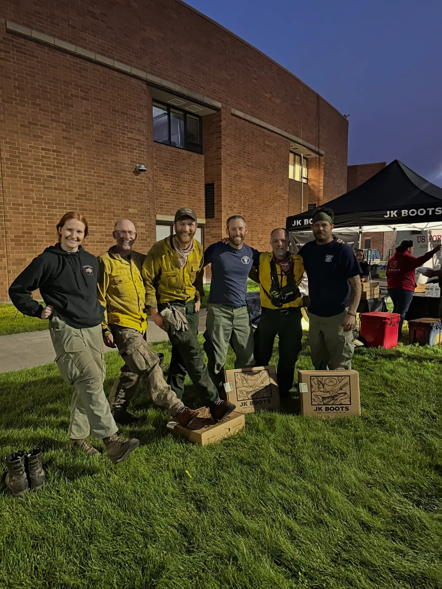 Six people stand on grass in front of a brick building, posing with cornhole boards labeled "JK BOOTS." A black tent and red equipment are visible in the background.